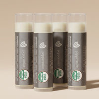 Four Beauty by Earth Organic Peppermint Lip Balms arranged upright in a staggered group, featuring gray packaging, translucent caps, and USDA Organic labels against a warm beige studio background.
