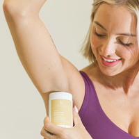 Woman applying beauty by earth deodorant to her armpit on a plain background