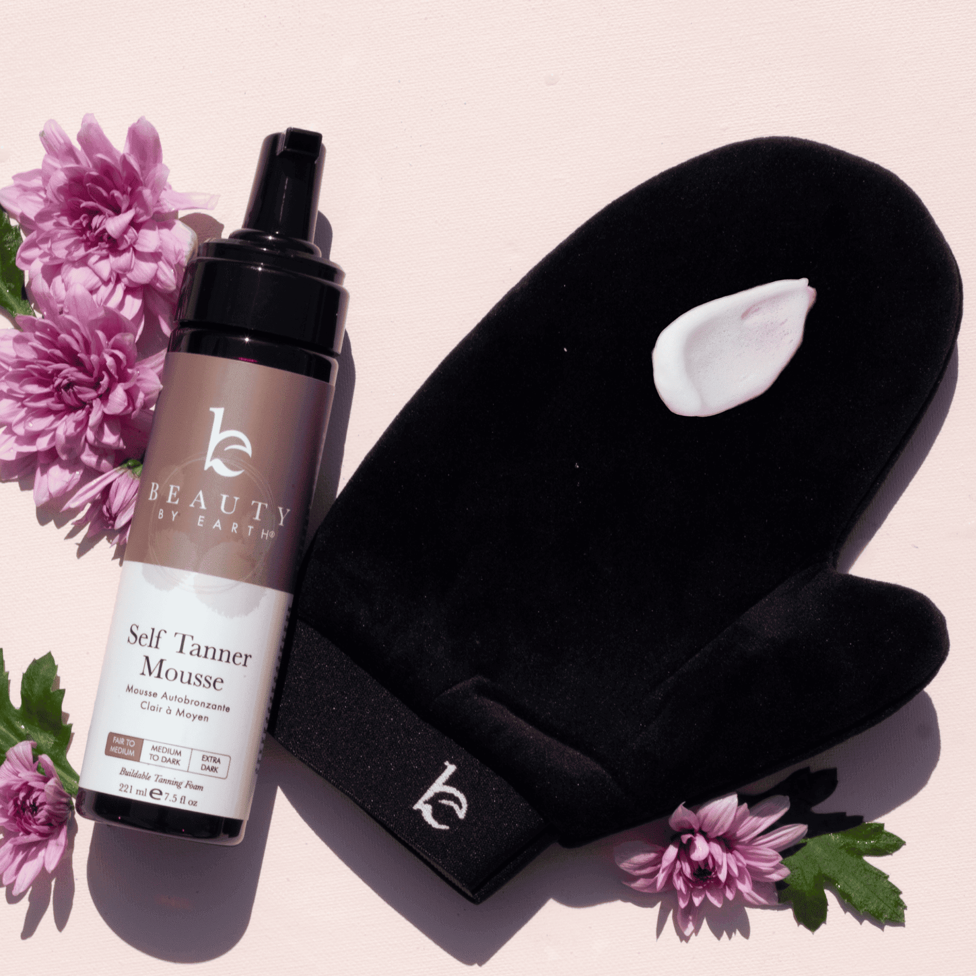 Beauty by Earthmott Self Tanner Mousse bottle with black mitt and pink flowers on a light pink background
