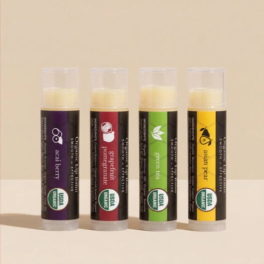 Four Beauty by Earth Organic Superfruit Lip Balms in Acai Berry, Grapefruit Pomegranate, Green Tea, and Asian Pear varieties, displayed upright in a row against a warm beige studio background. Each tube features colorful packaging and a USDA Organic certification label.