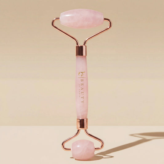 Beauty by Earth Rose Quartz Facial Roller featuring two natural pink quartz stones, rose-gold metal hardware, and a branded pink stone handle, standing upright against a warm beige background with soft directional shadows.