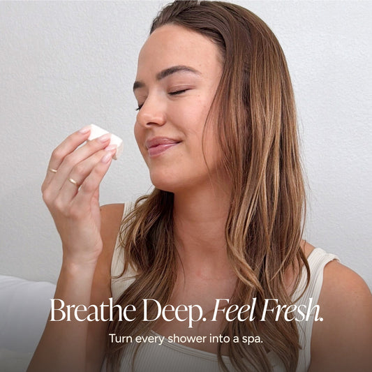 A woman with long brown hair closes her eyes while holding a shower steamer near her face, with the text 'Breathe Deep. Feel Fresh.