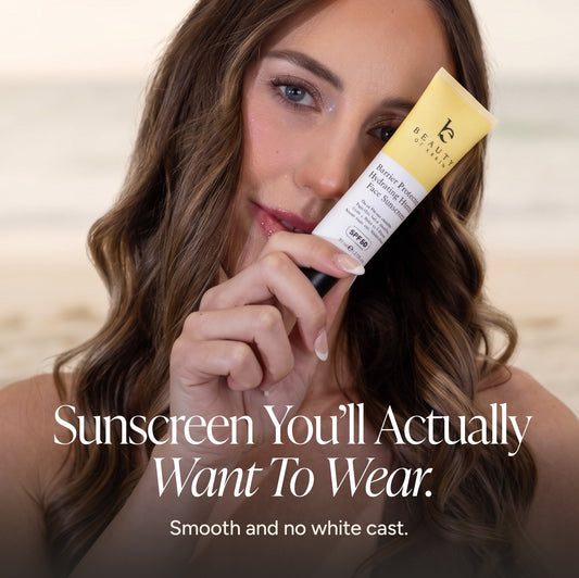 A woman holding a tube of Face Sunscreen against a blurred beach background, with text overlay reading 'Sunscreen You'll Actually Want To Wear. Smooth and no white cast.