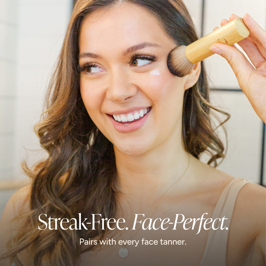 A smiling woman with long brown hair applies face tan using a kabuki brush with a bambii handle. Text at the bottom reads 'Streak-Free.