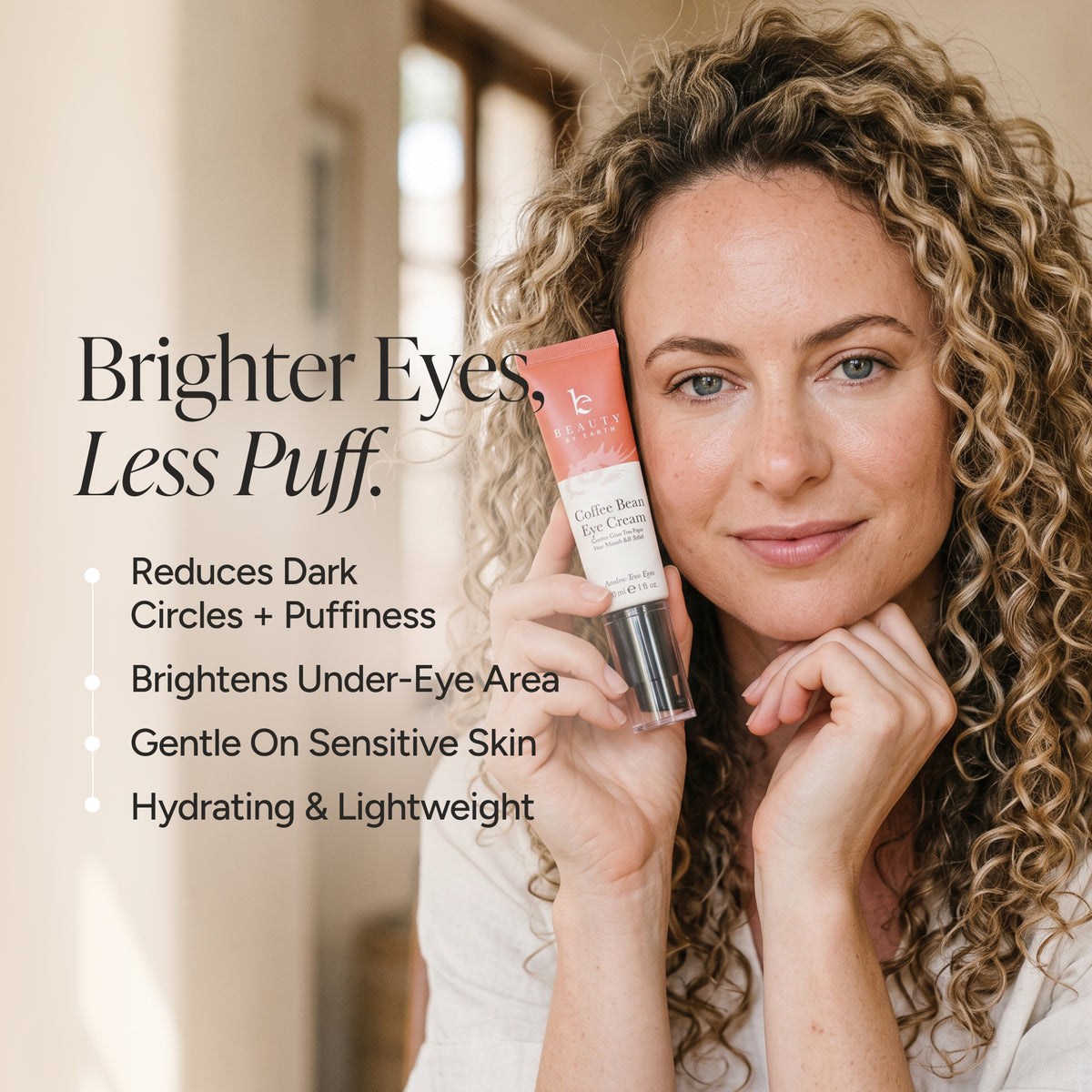 A woman with curly blonde hair holds a tube of eye cream next to text describing its benefits: 'Brighter Eyes, Less Puff.' The tube is peach and white with a black cap, and the background is a softly blurred indoor setting.