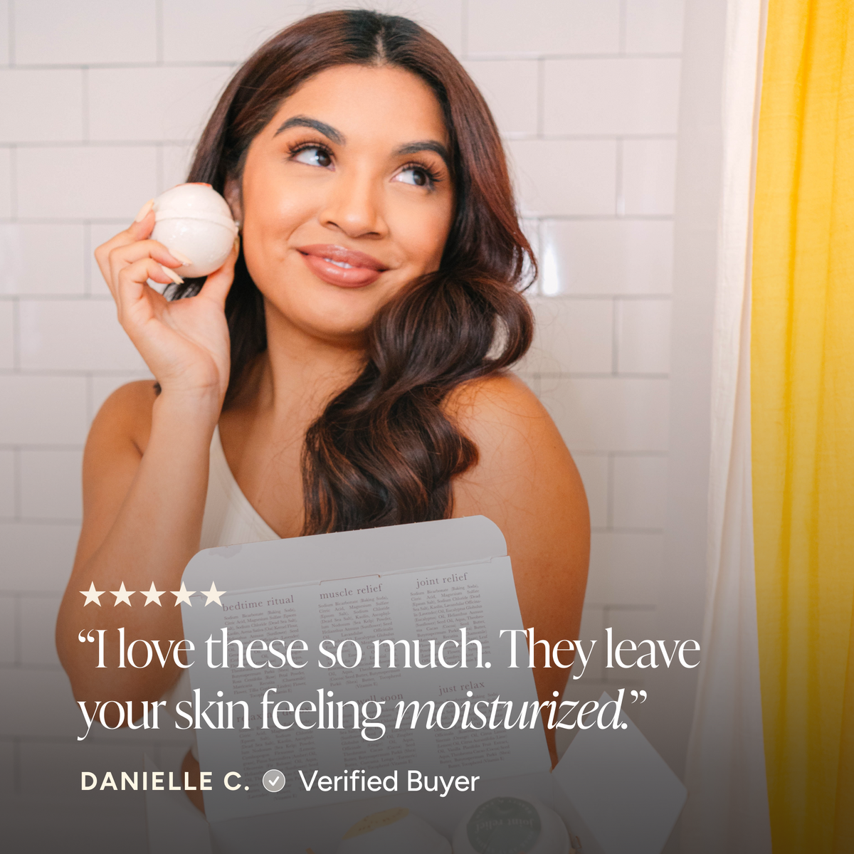 A woman in a bathroom holds a bath bomb near her face while looking upward with a smile. A translucent review card with a five-star rating and the text, 'I love these so much.