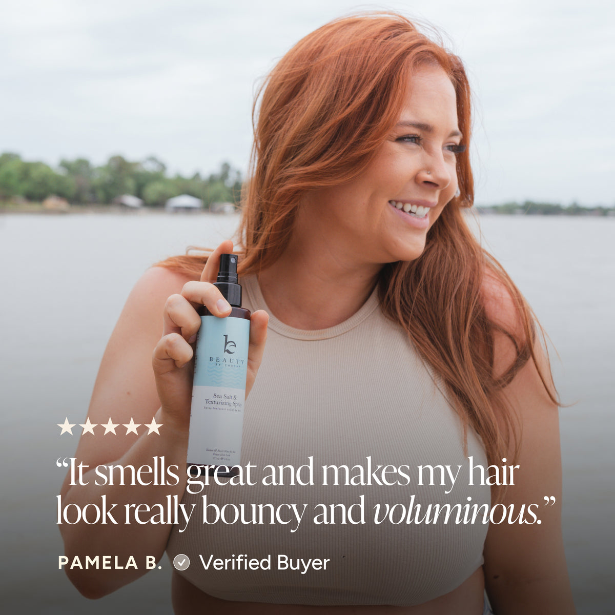 A woman with long red hair holds up a bottle of hair product outdoors near the water. The bottle is white with a black spray top and a pale blue label.