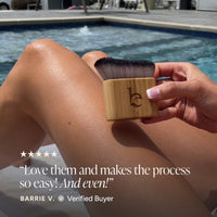 A hand holds a bamboo blending brush with dark bristles against a person's leg, with a swimming pool in the background and a review quote overlaid at the bottom.