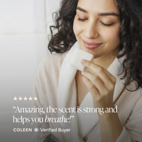 A woman in a white robe holds a small shower steamer near her face, with a soft-focus bathroom-like background and overlaid text reading a review: 'Amazing, the scent is strong and helps you breathe!' COOLEN Verified Buyer.