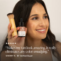 A woman holds a kabuki brush and a small bottle of tanner drops.