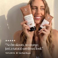 A smiling woman holds two tubes of Beauty by Earth Self Tanning Tanning Lotion against a warm sunlit wall.