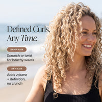 A smiling woman with long, defined curly blonde hair stands outdoors near the beach, wearing a black ribbed top. Text on the left reads, 'Defined Curls, Any Time.