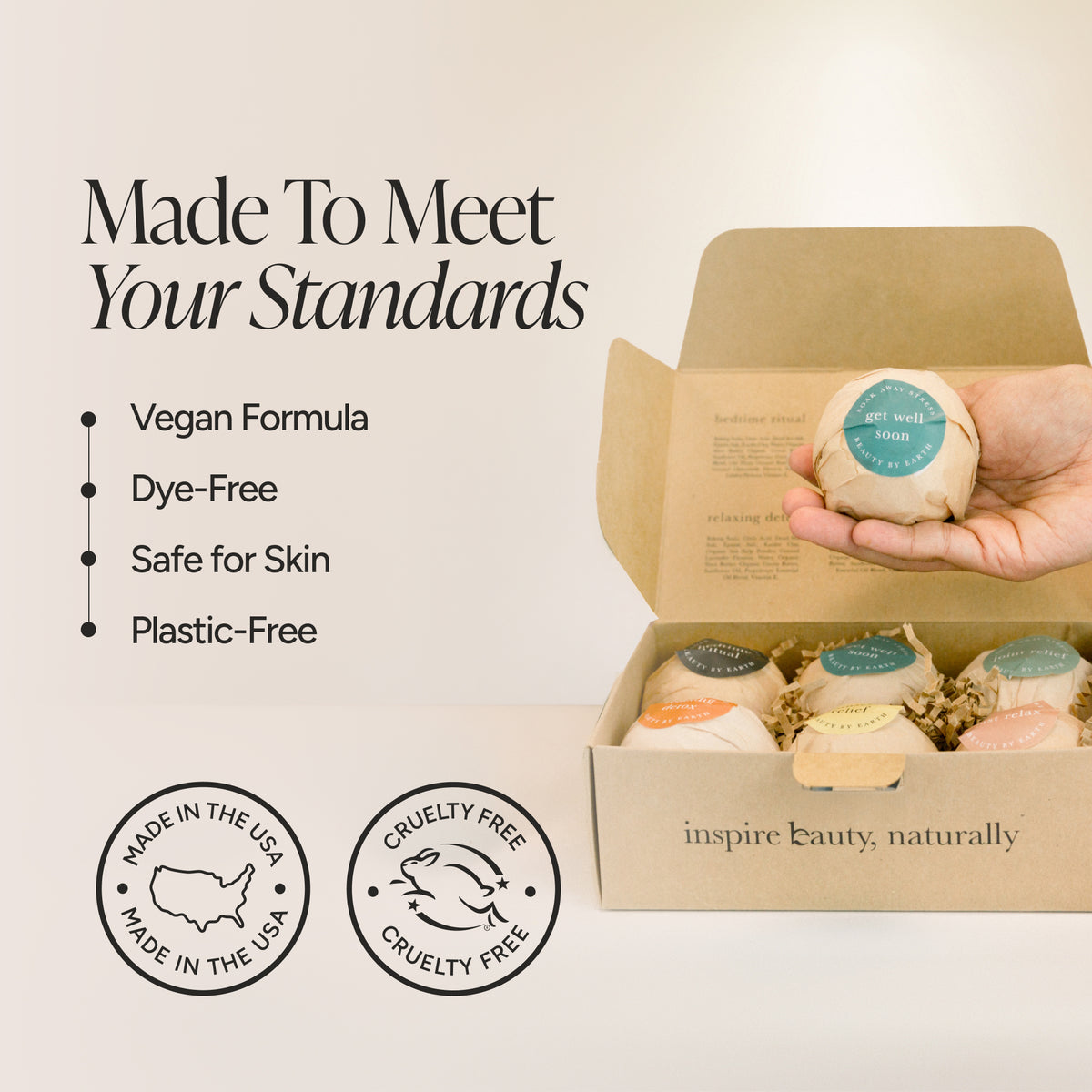 A hand holds a round bath bomb wrapped in beige paper above an open kraft gift box containing several bath bombs. Text on the left reads 'Made To Meet Your Standards' with bullet points for vegan formula, dye-free, safe for skin, and plastic-free, plus two circular badges for Made in the USA and Cruelty Free.