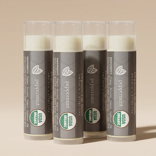 Four Beauty by Earth Organic Peppermint Lip Balms arranged upright in a staggered group, featuring gray packaging, translucent caps, and USDA Organic labels against a warm beige studio background.