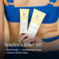 A close-up lifestyle shot of a woman holding two tubes of Barrier Protection Mineral Sunscreen SPF 50. The woman is wearing a bright blue knit top, and the background is a warm beige tone.