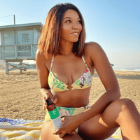 Woman in a bikini sitting on the beach with a bottle of sunscreen.