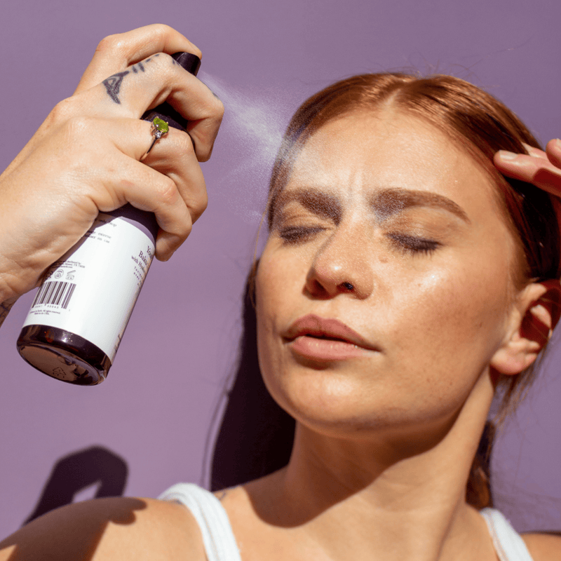 Person applying a product to their face with a spray bottle against a purple background