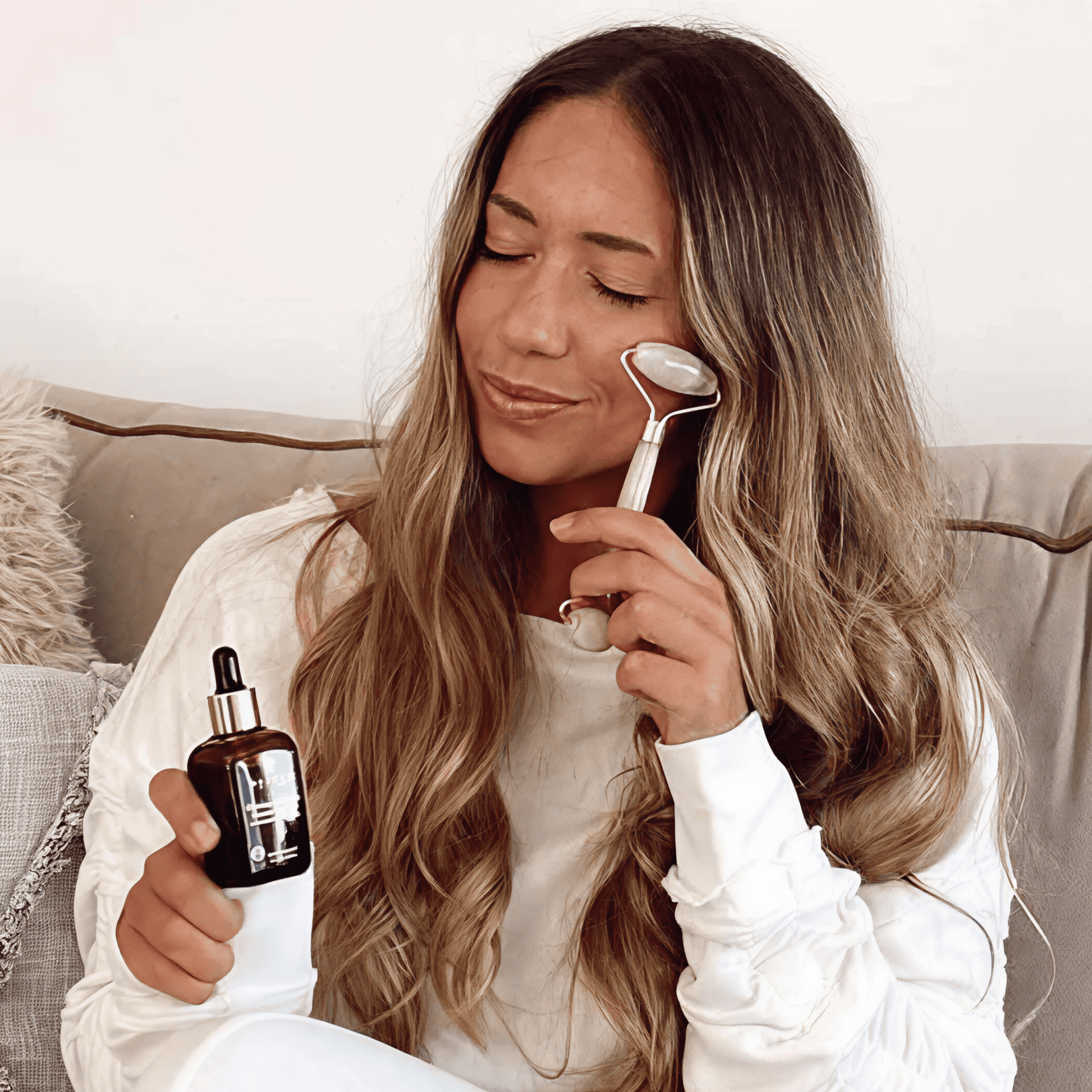 Woman applying skincare product with a roller and dropper bottle on a couch.
