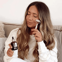 Woman applying skincare product with a roller and dropper bottle on a couch.