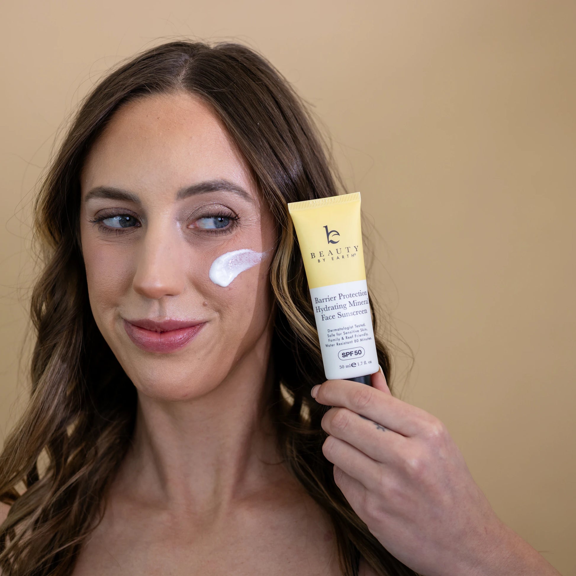 Woman applying sunscreen to her face with a tube of sunscreen in front of a beige background