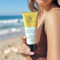 Person holding a tube of 'Beauty by Earth' sunscreen on a beach.
