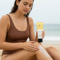 Woman applying sunscreen on her arm with a tube of sunscreen in her hand, beach setting.