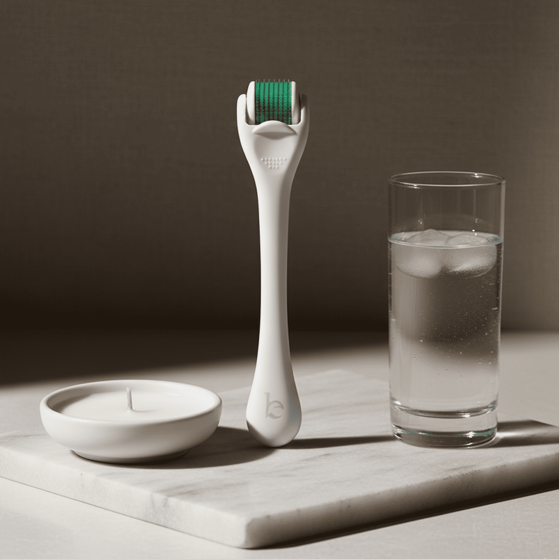 White facial derma roller with green head on a marble surface next to a glass of water and candle