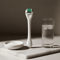 White facial derma roller with green head on a marble surface next to a glass of water and candle