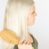 Woman with long blonde hair using a wooden paddle brush against a light background