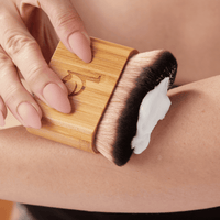 Person using a wooden brush with bristles on a person's arm, applying a white cream or lotion.