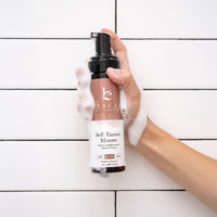 Hand holding a bottle of Beauty by Earth self Tanner Mousse against a white tiled background