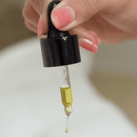 Hand holding a dropper with nail and cuticle oil yellow liquid against a neutral background