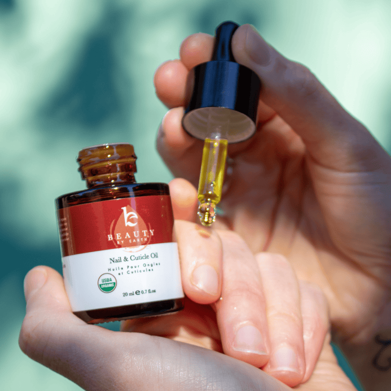 Hand holding a bottle of Beauty by Earth nail and cuticle oil with a dropper filled with yellow liquid.