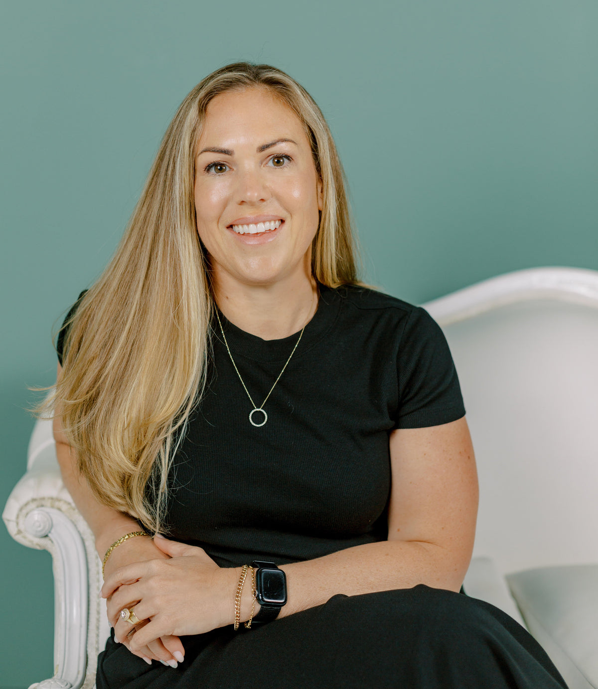 Woman sitting on a white chair wearing a black dress against a teal background