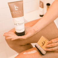 A model holding self tanning body lotion in one hand and kabuki body blending brush in the other while white lotion is applied a bit on a leg.