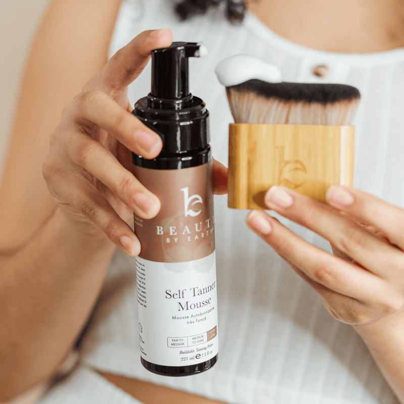 Person holding a bottle of 'Beauty by Earth' self-tanning mousse with a brush.