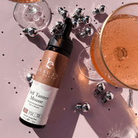 Self Tanner Mousse bottle with champagne glasses on a pink background