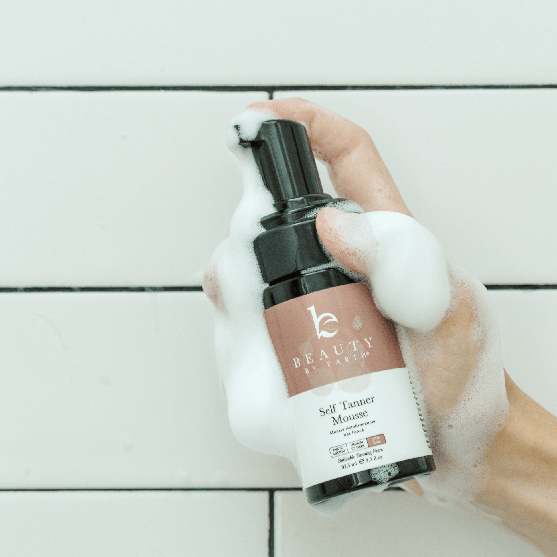 Hand holding a bottle of Beauty by earth Self Tanner Mousse on a light wooden surface