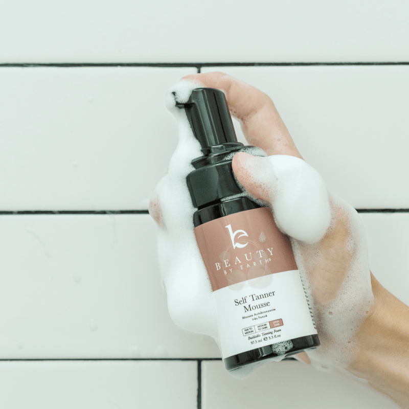 Hand holding a bottle of Beauty by earth Self Tanner Mousse on a light wooden surface