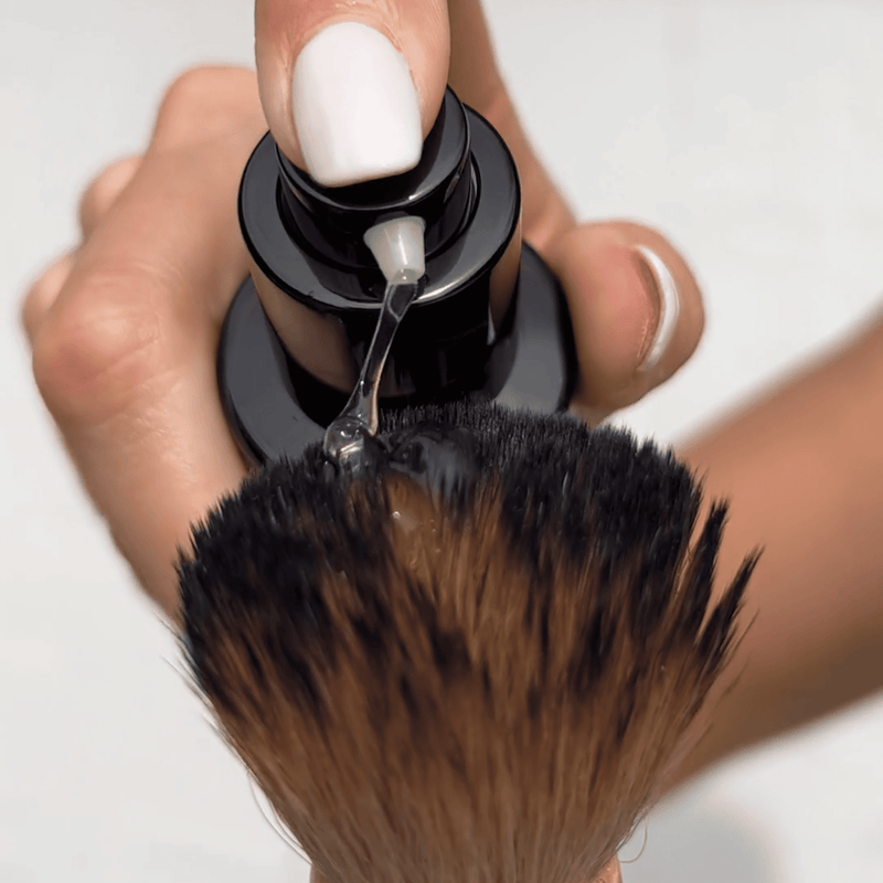 Hand holding a kabuki facial brush with a close-up of the self tanning facial serum texture on the brush