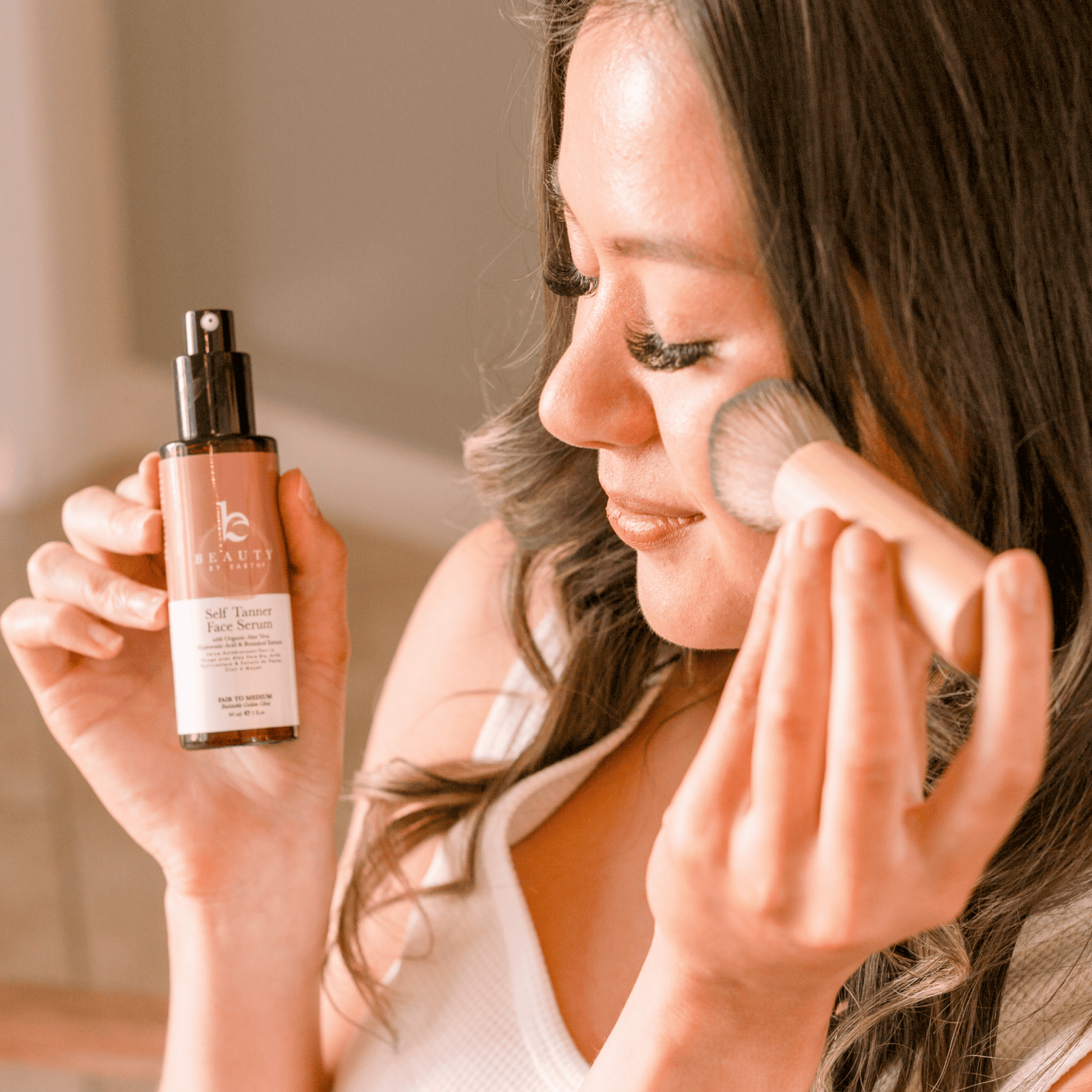 Woman applying self tanning facial serum with a kabuki facial brush and holding a bottle of Beauty by Earth self tanning facial serum in hand.