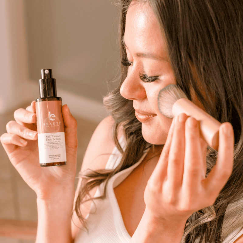 Woman applying self tanning facial serum with a kabuki facial brush and holding a bottle of Beauty by Earth self tanning facial serum in hand.