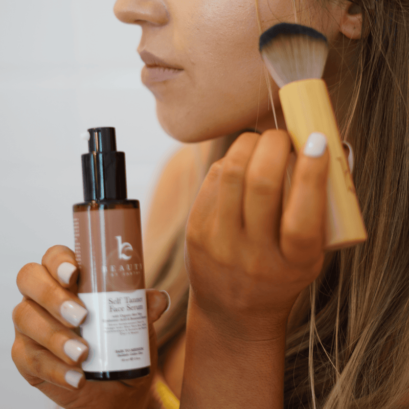 Person holding a bottle of Beauty by Earth self tanning serum and a kabuki facial brush against a neutral background