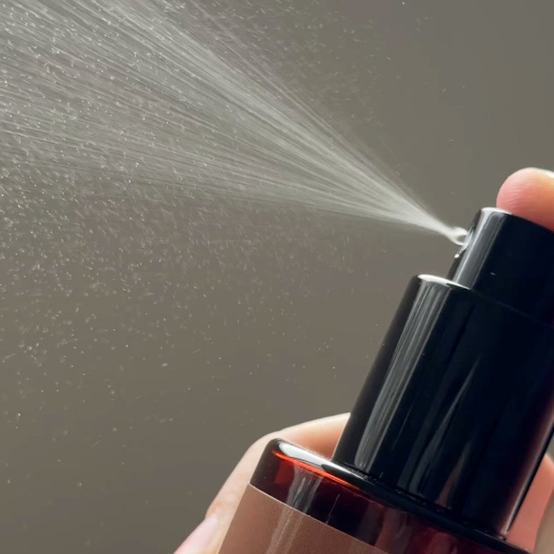 Hand holding a water bronzing face mist being released against a gray background