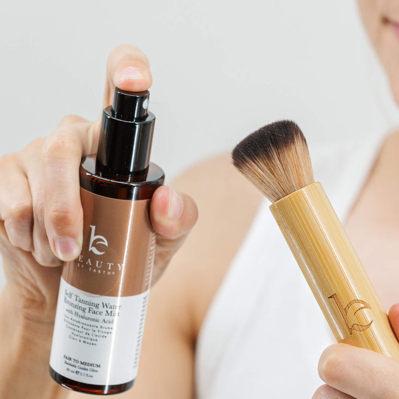 Hand holding a bottle of 'beauty by earth' tanning water bronzing mist and a kabuki brush with a blurred background