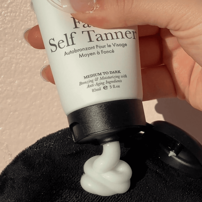 Hand holding a tube of Face Self Tanner with visible texture white lotion on a black applicator. 