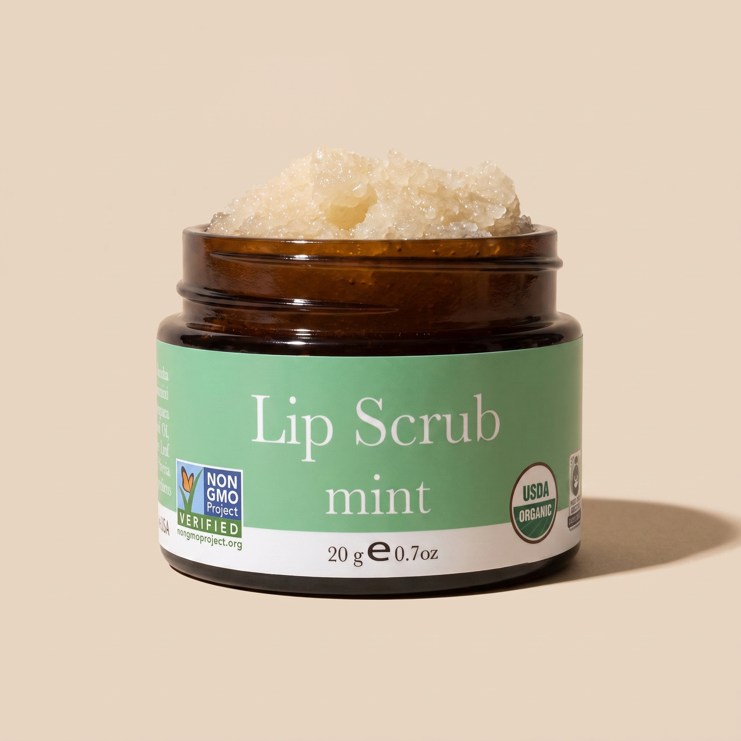 An open amber glass jar of lip scrub with a green label reading 'Lip Scrub mint' on a warm beige background. The scrub is visible at the top of the jar, and certification logos for Non-GMO Project and USDA Organic appear on the label.