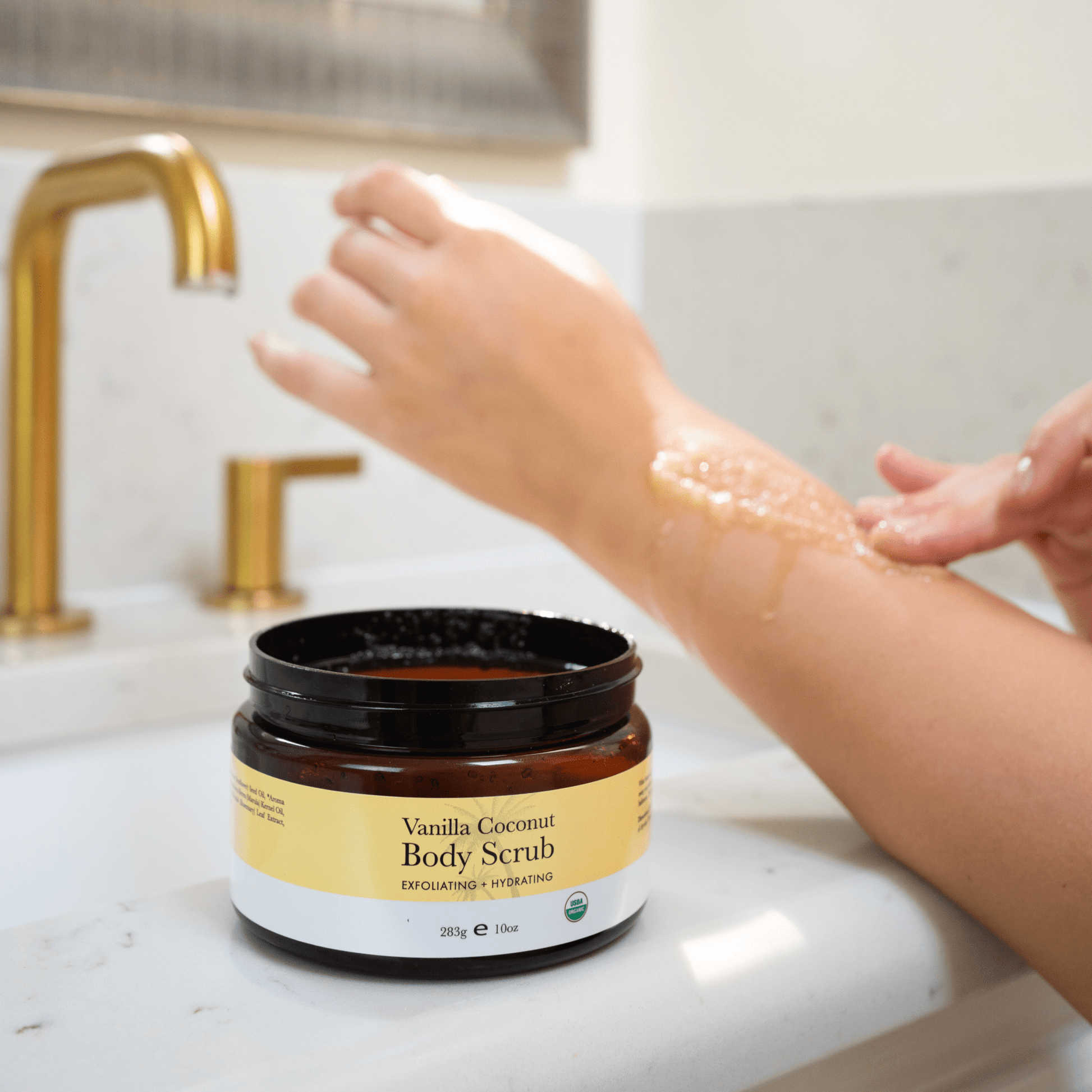 Vanilla Coconut Body Scrub jar on a bathroom counter with a hand applying scrub.