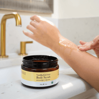 Vanilla Coconut Body Scrub jar on a bathroom counter with a hand applying scrub.