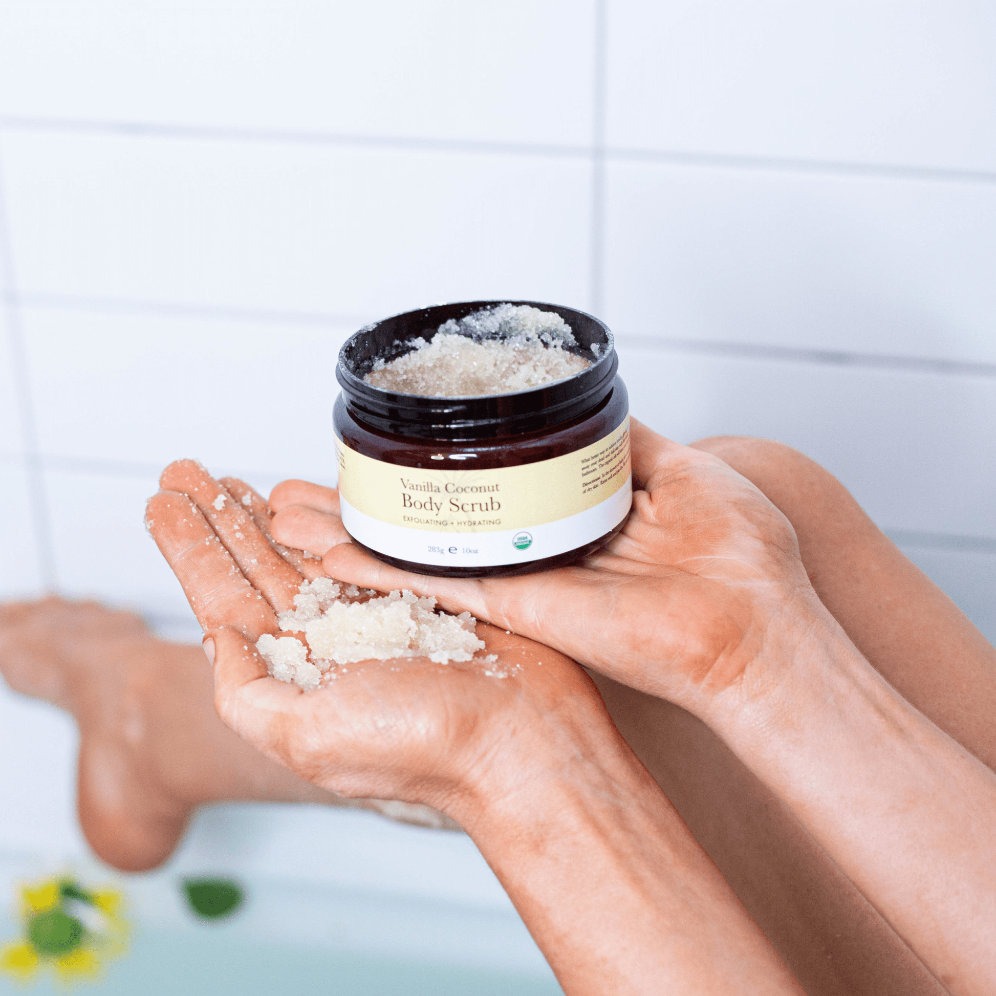 Person holding a jar of Vanilla Coconut Body Scrub with visible scrub in hands against a blurred bathroom background.